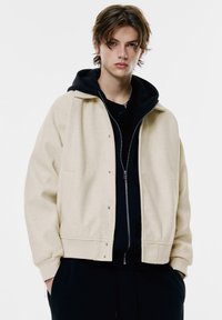 Beige uld bomberjakke med knaplukning og ribbede manchetter, lagd over en sort zip-up hoodie med en frontlomme.