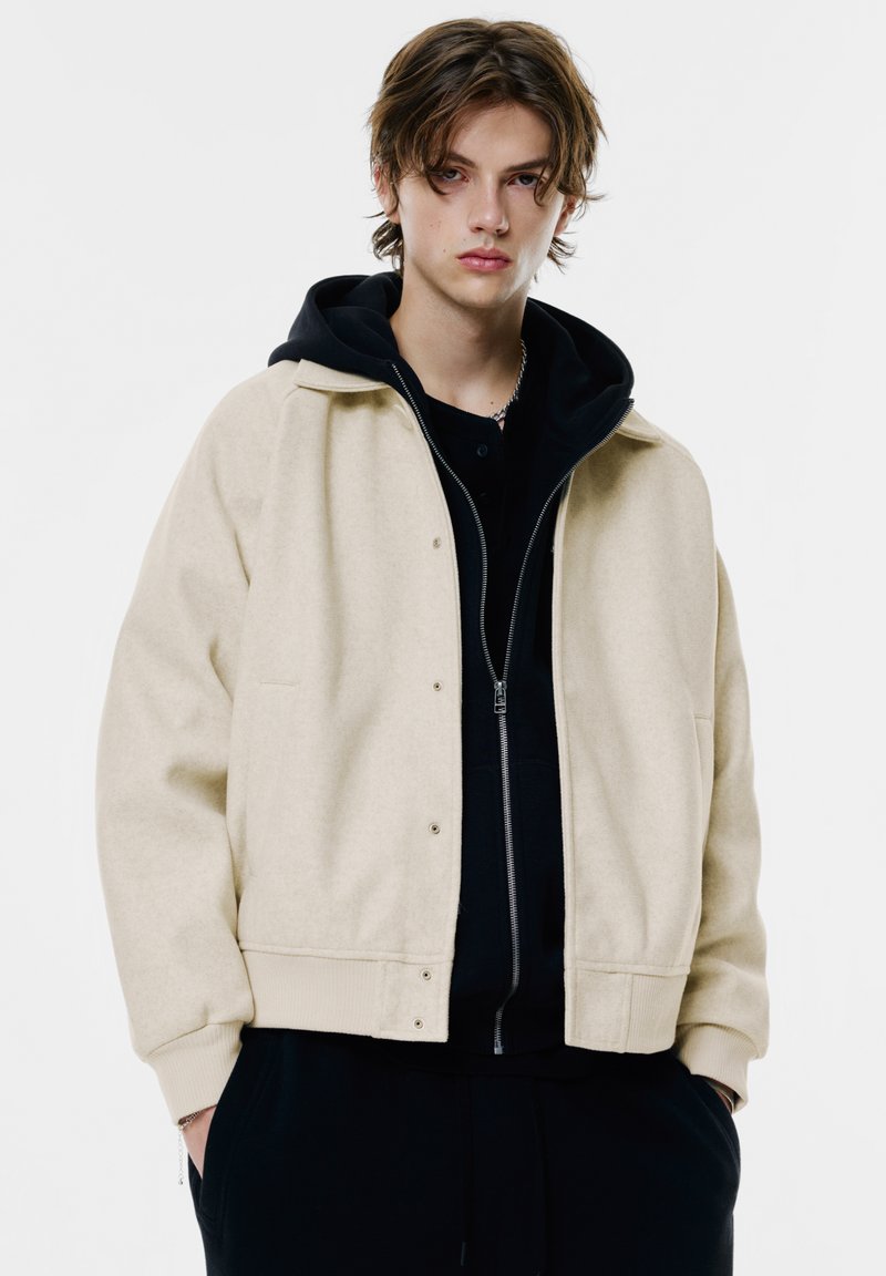 Beige uld bomberjakke med knaplukning og ribbede manchetter, lagd over en sort zip-up hoodie med en frontlomme.