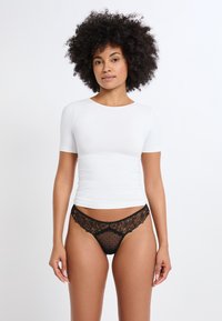 Top blanco de manga corta con un diseño ajustado, combinado con un tanga negro de encaje con lunares y acentos florales. Textura suave en general.