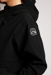 Zwarte waterdichte jas met capuchon, beschikt over een logopatch op de linkermouw, heeft een soepele textuur en een losse, ontspannen pasvorm.