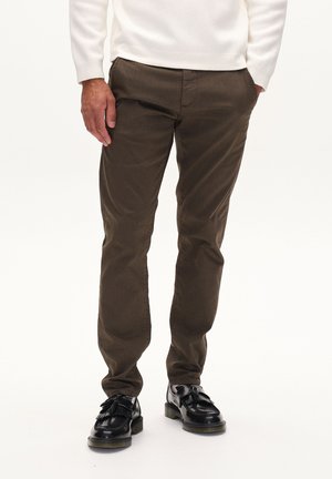 Mann trägt braune Cordhose, schwarze Tasselloafer und ein weißes langärmeliges Oberteil, steht mit Händen nahe den Taschen.