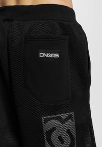 Čierne mriežkové joggers nohavice majú textúrovanú látku, pevnú čiernu kapsu s logom a kontrastný grafický dizajn na boku.