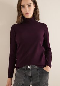 Burgundowy sweter z prążkowanym kołnierzem, z długimi rękawami i lekko dopasowanym krojem, w połączeniu z szarymi dżinsami z kieszeniami i widocznym szwem.