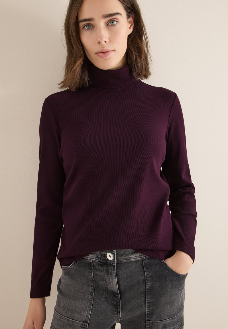 Burgundowy sweter z prążkowanym kołnierzem, z długimi rękawami i lekko dopasowanym krojem, w połączeniu z szarymi dżinsami z kieszeniami i widocznym szwem.