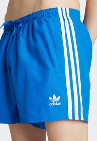 Kék rövidnadrág fehér háromcsíkos dizájnnal, elasztikus derékkal és állítható zsinórral. Hímzett Adidas logóval rendelkezik.