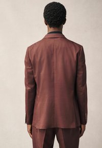 Chaqueta de traje a medida en color burdeos con un diseño elegante, detalle de abertura en la parte posterior y mangas largas. Textura de tela suave y ajuste estructurado.