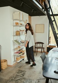 Un pull gris surdimensionné et un pantalon noir plissé. Une étagère blanche porte des décorations en bois, des livres et une lampe ronde. La lumière naturelle éclaire la scène.