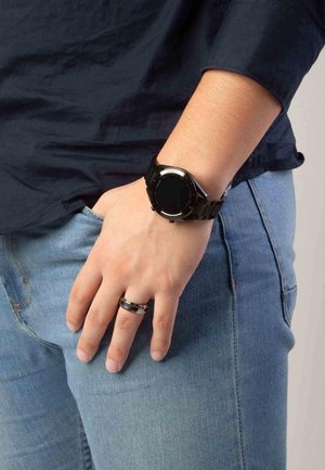 Black digital watch with a round face, metallic accents, and a textured band, worn on a hand placed in denim pocket. Silver ring on the other hand.