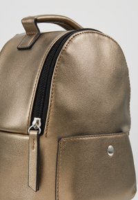 Metallisch goldener Rucksack mit runder Form, glatter Textur, kontrastierendem schwarzen Reißverschluss und einer Fronttasche mit Druckknopfdetails.
