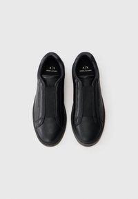 Um par de ténis slip-on pretos da Armani Exchange com faixas elásticas na parte superior, exibidos sobre um fundo cinza claro liso.