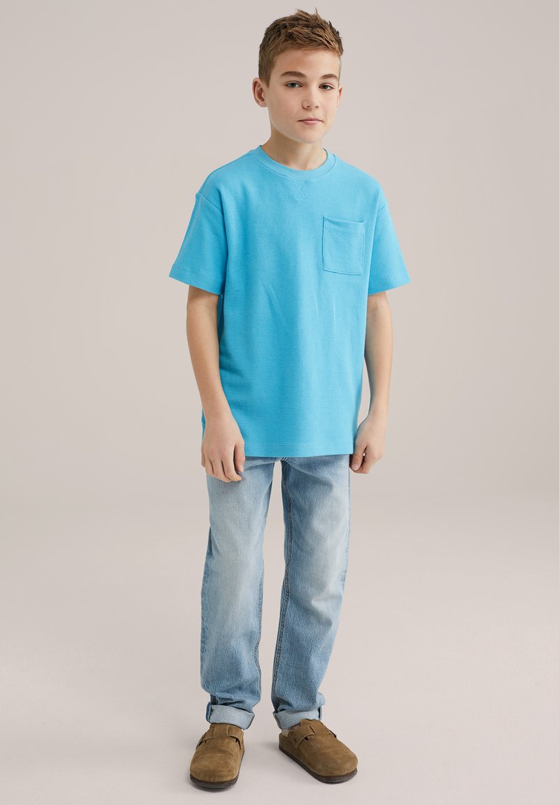 Svijetloplavi majica s kratkim rukavima s džepom na prsima, u kombinaciji s izblijedjelim trapericama i smeškim cipelama bez pertli. Jednostavan dizajn, opušteni kroj.