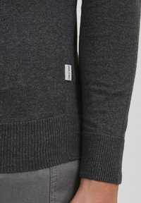 Dunkelgrauer Strickpullover mit Ärmeln und Saum, an dem ein kleines "G-Blend"-Textiletikett in der Nähe der Seitennaht über grauen Hosen sichtbar ist.
