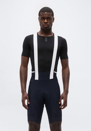 Homme portant un maillot de cyclisme noir à manches courtes et un cuissard noir avec des bretelles blanches, debout face à l’avant sur un fond blanc.