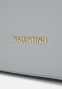 Szürke, texturált bőr felület, amelyen egy arany dombornyomott "VALENTINO" logo látható, kiemelve az elegáns dizájnt és a prémium anyagot.