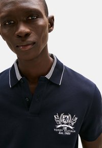 Polo azul marino con un logo bordado en blanco que incluye banderas, cuello a rayas y tapeta con tres botones. Tejido texturizado, ajuste clásico.