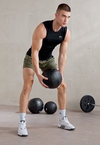 Fitness equipment: black medicine ball, smooth texture. Worn by a person in a sleeveless black top and olive green shorts, white athletic shoes.