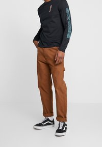 Hombre vistiendo pantalones marrones arremangados, camisa negra de manga larga con texto azul en las mangas, calcetines blancos y zapatillas negras de corte bajo sobre un fondo liso.