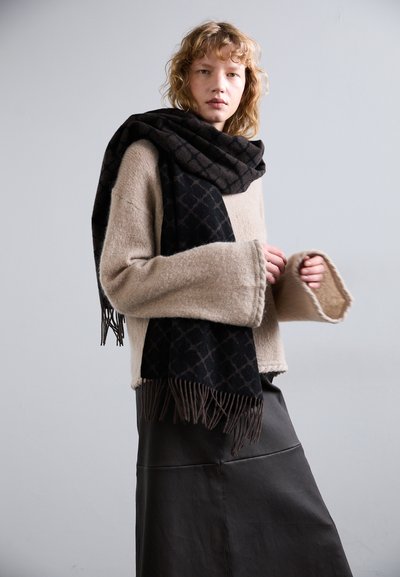 Pulover bej, călduros, din tricot cu mâneci largi. Șal negru cu un model geometric și capete franjurate. Fustă neagră din piele texturată.