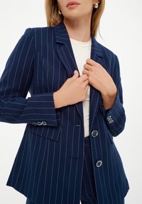 Blazer de rayas azul marino con un ajuste entallado, solapas con muesca, dos botones y un brillo sutil, combinado con una blusa de color claro.
