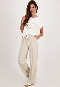 Vrouw die glimlacht, in een witte T-shirt met opgerolde mouwen en de tekst "Atelier Studio", beige trekkoordbroek en witte sneakers, staande op een witte achtergrond.