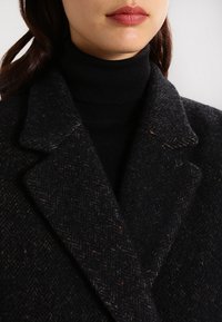 Donna che indossa un cappotto di lana nera strutturato sopra un maglione nero a collo alto, con un leggero rossetto rosa sulle labbra.
