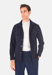 Giacca di lana blu navy con colletto, due tasche sul petto e chiusura a bottoni. Texture liscia e vestibilità aderente. Indossata sopra un top grigio chiaro.