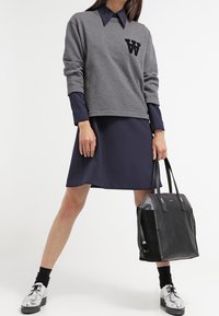 Harmaa huppari, jossa on musta "W"-logo, käytetty tummansinisen mekon päällä. Musta käsilaukku ja kiiltävät hopeakengät täydentävät asukokonaisuutta. Mustat sukat näkyvät.