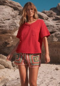 T-shirt rouge à manches courtes avec col en V et poche poitrine, associé à un short multicolore à motifs de carrés verts, rouges et blancs.