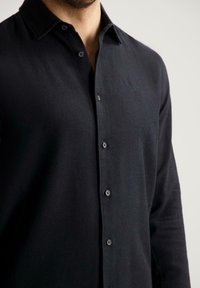 Man in een zwart button-up overhemd met lange mouwen en een klein geborduurd logo op de linkerborst tegen een effen achtergrond.