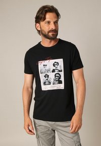Homme avec une barbe portant un t-shirt noir présentant quatre squelettes portant des chapeaux et des lunettes de soleil avec le texte "Voyage à Vegas" et "Créez des souvenirs".