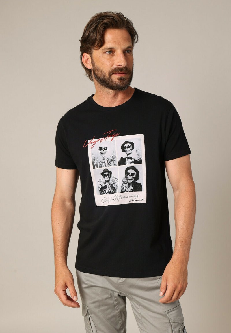 Homme avec une barbe portant un t-shirt noir présentant quatre squelettes portant des chapeaux et des lunettes de soleil avec le texte "Voyage à Vegas" et "Créez des souvenirs".