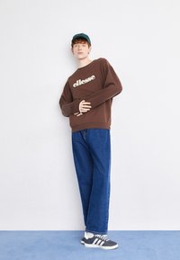 Sudadera marrón con el logotipo de "ellesse", jeans de mezclilla azul y zapatillas de navy con rayas blancas. El modelo lleva una gorra de color teal. Texturas suaves, ajuste casual.