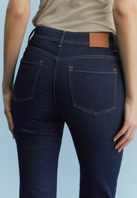 Osoba v tmavě modrých džínech a béžové košili, zobrazená od pasu po střed stehna, na světle modrém pozadí.