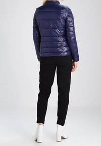 Marinblå pufferjacka med slim fit, horisontell quiltad design, ståkrage och glänsande yta, i kombination med svarta byxor och vita klackstövlar.