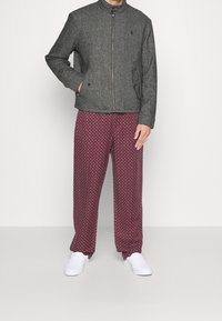 Grå zip-up jacka med ståkrage och sidofickor, ihop med vinröda mönstrade byxor. Modellen bär vita slip-on skor.