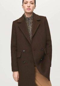 Cappotto doppiopetto in misto lana marrone con grandi bottoni neri, rever con risvolto e tasche anteriori inclinate. Si intravede un maglione a fantasia.