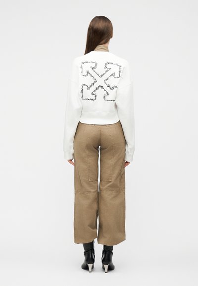 Femme portant un sweat-shirt blanc avec un motif de flèche noire dans le dos, un pantalon beige à jambes larges avec des détails en patch, et des bottines à talons noires.