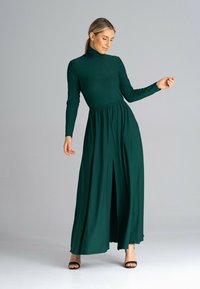 Zielona, długa sukienka maxi z długimi rękawami, wysokim dekoltem, marszczoną talią i rozcięciem z przodu, wykonana z miękkiego, zwiewnego materiału. Czarne sandały na obcasie z odkrytymi palcami.