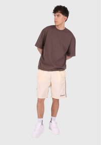 Braunes Baumwoll-T-Shirt, beigefarbene Shorts mit seitlichen Streifen und weiße Sneaker. Lässige Passform, kurze Ärmel und bestickte Details an den Shorts.