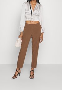 Brązowe, dopasowane spodnie w zestawieniu z satynowym, srebrnym topem odsłaniającym brzuch; modelka trzyma kremową, teksturowaną kopertówkę i nosi czarne, sandały na wysokim obcasie ze sznurowaniem.