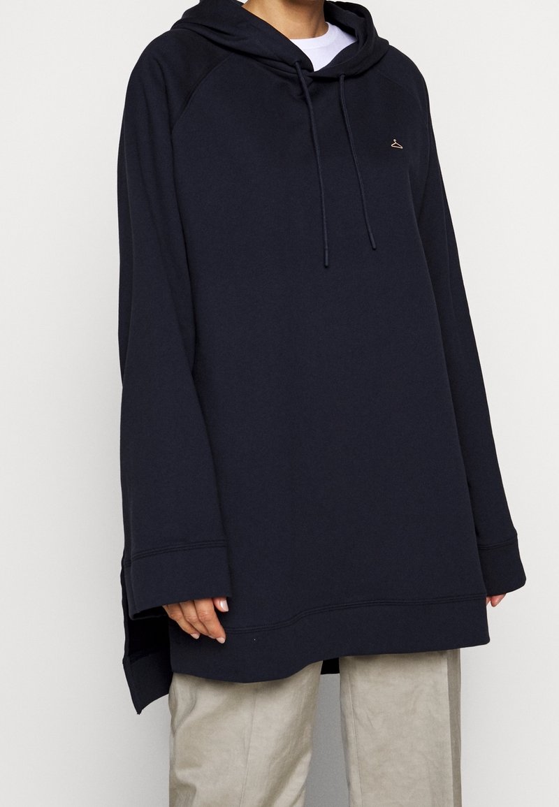 Personne portant un sweat à capuche noir oversize avec cordons et petit logo cintre, associée à un pantalon beige clair, debout devant un fond blanc.