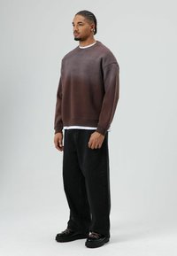 Bruine sweat met verloopkleur en een afgeronde halslijn, voorzien van kleine oogjesdetails, gecombineerd met losse zwarte broek en zwarte schoenen met roze veters.