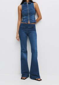Crop top en denim avec fermeture éclair et col, associé à un pantalon en denim évasé. Les deux sont dans une teinte bleue moyenne avec de subtils détails de couture.