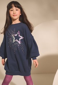 Abito felpa oversize blu navy con maniche a sbuffo, caratterizzato da un design a stella scintillante in paillettes. Abbinato a collant viola.