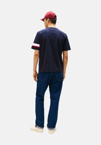 Camiseta azul marino de manga corta con franjas blancas y rojas en los puños. Combinada con jeans azul oscuro y una gorra roja.