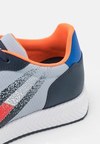 Scarpa sportiva grigia con tomaia testurizzata, colletto in rete arancione, tallone blu e suola bianca a zigzag. Presenta strisce diagonali colorate.