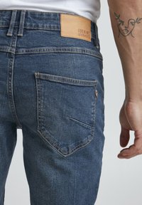 Temno modri kavbojke iz denima, ki imajo usnjeno oznako z napisom "DENIM JOURNEY", dizajn zadnjega žepa in diskretne šive.