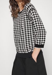 Pull en tricot à motif pied-de-poule noir et blanc avec manches trois-quarts et ourlet côtelé noir, doté d'un col rond.