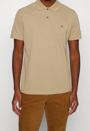 Beige Poloshirt aus Baumwolle mit klassischem Kragen, zwei Knöpfen, kurzen Ärmeln und kleinem grünen Logo auf der Brust. Getragen mit Baumwollcordhosen.