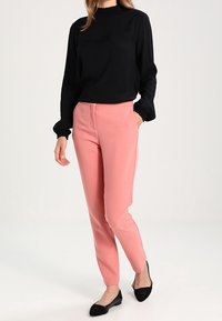 Blouse noire à manches longues avec encolure froncée et poignets à volants, assortie à un pantalon tailleur rose clair et des chaussures plates noires.